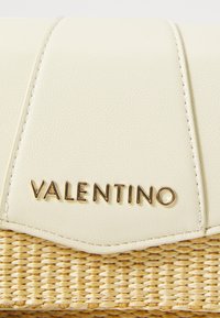 Bolsa de couro creme com base de palha trançada. Apresenta o logótipo em tom dourado "Valentino" e detalhes acolchoados. Textura suave e forma estruturada.