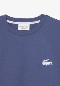 T-shirt girocollo in cotone blu con un piccolo logo di coccodrillo bianco sul lato sinistro del petto e un'etichetta al collo con la scritta "LACOSTE LOUNGEWEAR."