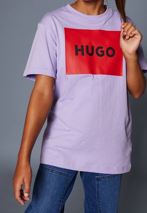 Persona che indossa una maglietta viola chiaro con un grande quadrato rosso con la scritta "HUGO" in lettere nere, abbinata a jeans blu.