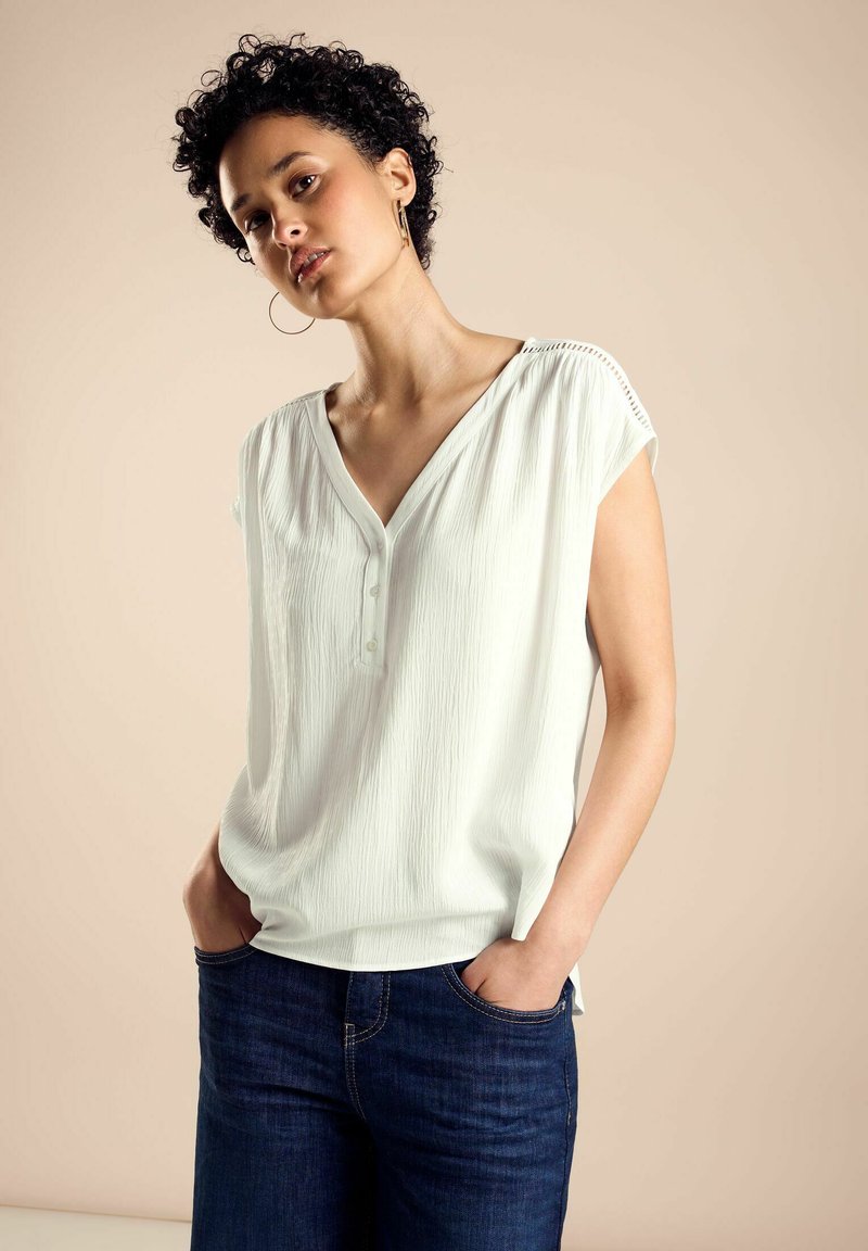 Weiße, strukturierte Bluse mit V-Ausschnitt und kurzen Ärmeln; kombiniert mit dunkelblauen Jeans. Das Design umfasst einen Knopfdetail und seitliche Schlitze.