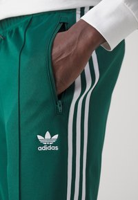 Zelené sportovní kalhoty z hladké látky, s bílými trojpruhovými akcenty, kapsou na zip a bílým logem Adidas.