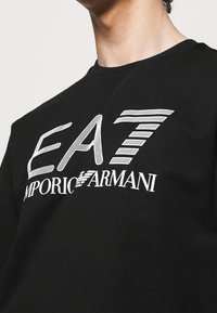 Närbild av en person som bär en svart tröja med vit "EA7 Emporio Armani"-logotyp tryckt på bröstet.