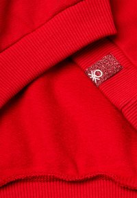 Felpa rossa con scollo e polsini a coste. Include una toppa glitterata testurizzata con un dettaglio bianco. Tessuto interno morbido.