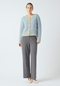 Vaaleansininen neulecardigan, jossa on beige reunus, nappikiinnitys, kaksi etutaskua ja harmaat suorat housut.