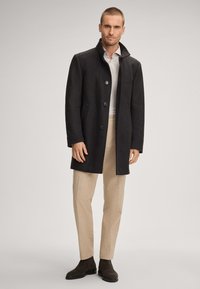 Mann in einem dunkelgrauen Knopfmantel, weißem Hemd, beigen Hosen und dunkelbraunen Stiefeln, der vor einem hellgrauen Hintergrund steht.