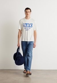 Hvit t-skjorte med blå "HUGO CREATIVE DEPT." grafikk, blå jeans, brune flip-flops og en marineblå duffelbag. Nøytral bakgrunn.