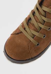 Brown suede ankle boot with olive green laces, round toe, metal eyelets, and textured surface. Features reinforced stitching and rubber sole.