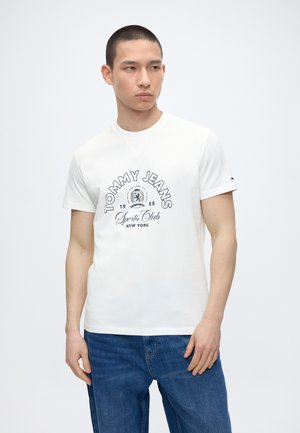 Ung mand med kort hår iført en hvid Tommy Jeans Sports Club T-shirt og blå jeans, der står foran en ensartet lys baggrund.