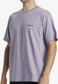 Camiseta de algodón lavanda con cuello redondo, mangas cortas y un logotipo gráfico en diseño circular en el pecho.