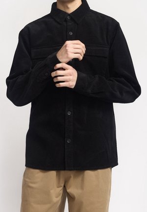 Homme boutonnant le poignet d'une chemise noire en velours côtelé à manches longues, portant un pantalon beige, sur un fond clair uni.