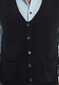 Svart stickad väst med v-ringning, sex knappar, två framfickor och ribbad textur, bärs över en ljusblå skjorta.