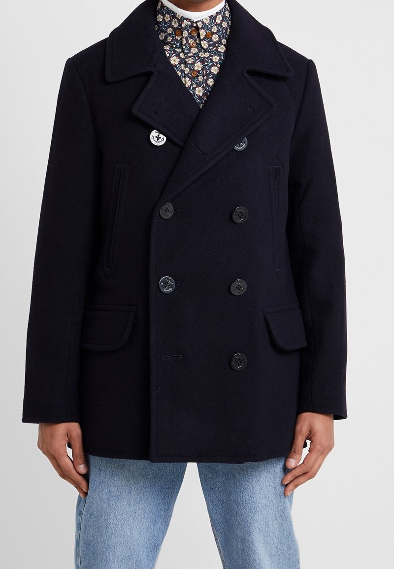 Cappotto peacoat in lana blu navy con chiusura a doppio petto, ampi risvolti, tasche laterali e vestibilità sartoriale, indossato sopra una camicia a fantasia.