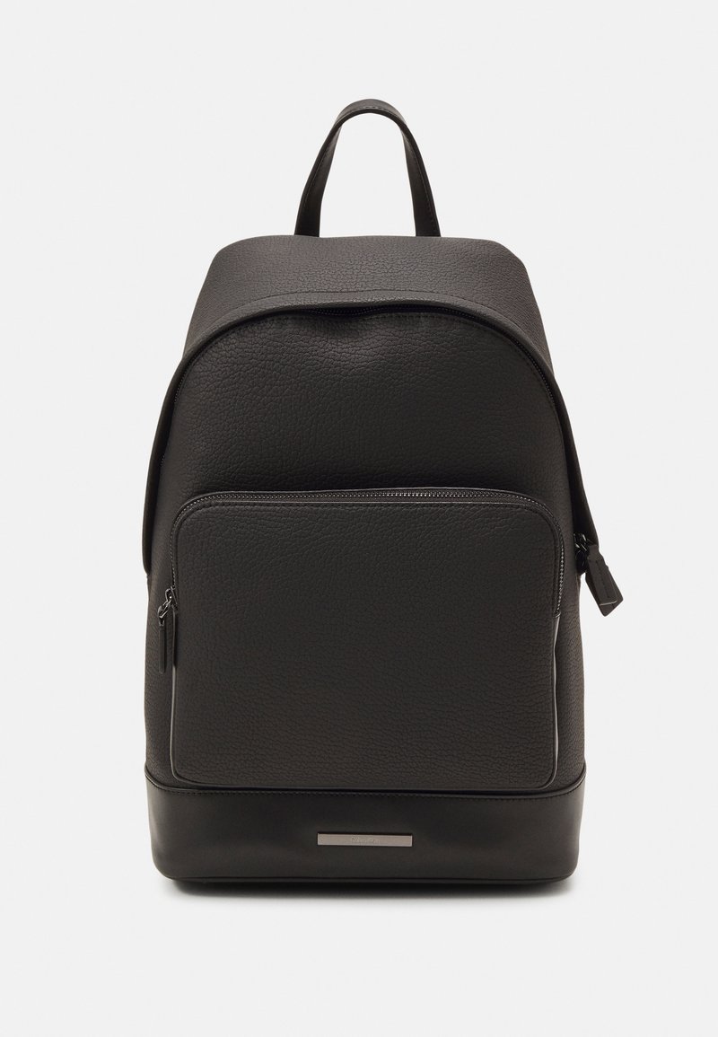 Zaino in pelle nera con superficie texture, parte superiore arrotondata, tasca frontale con zip e un dettaglio metallico alla base. Spallacci regolabili non visibili.