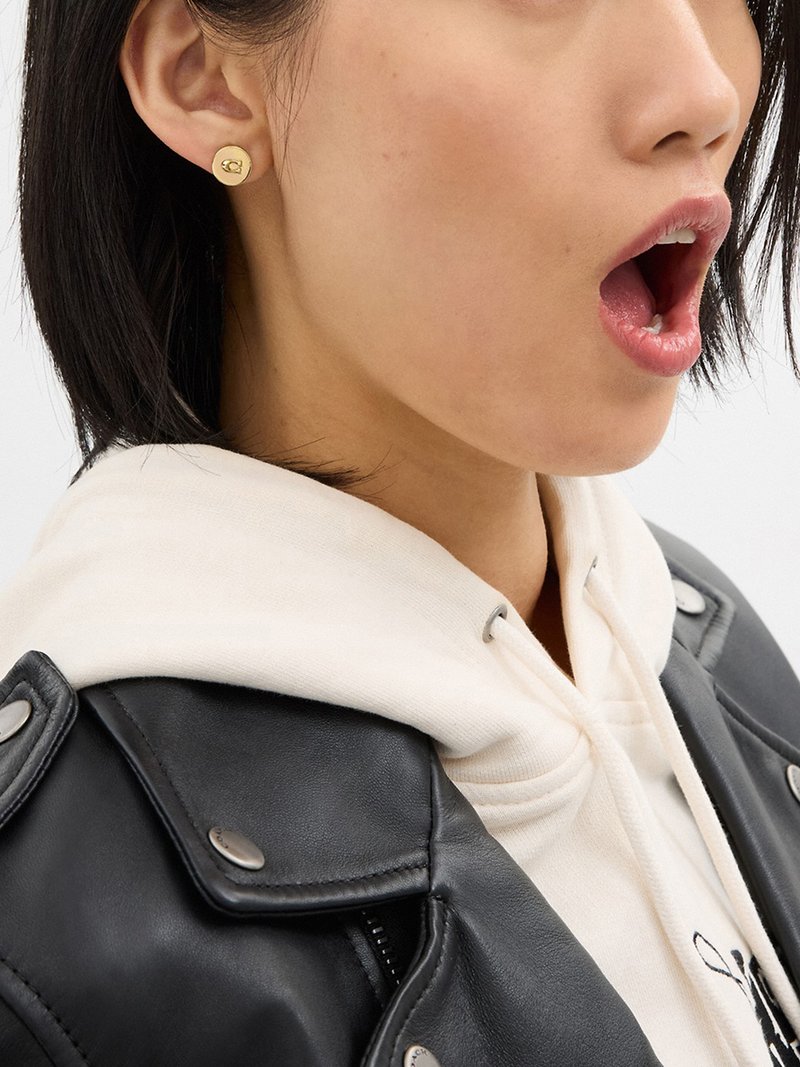 Woman with short black hair wearing gold round stud earring, white hoodie, and black leather jacket, mouth open as if speaking or singing.