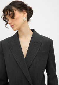 Blazer a righe grigio scuro con scollatura a V profonda, rever affilati e una sola tasca sul petto. Presenta un tessuto liscio e un design strutturato.