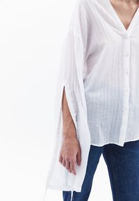 Weißes Hemd aus leichtem Stoff, mit langen Ärmeln und seitlichen Schlitzen sowie einem strukturierten Muster. Kombiniert mit blauen Jeans.
