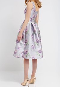 Hihaton, laventelinvärinen mekko, jossa on kukkakuosi ja liehuva helma. Valmistettu kiiltävästä kankaasta, jossa on nauhaakseli takana.