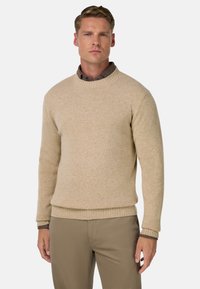 Béžový svetr s kulatým výstřihem z měkké pletené látky, s žebrovanými manžetami a lemem. Nošený přes tmavou košili s límečkem, doplněný khaki kalhotami.