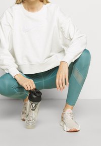 Sudadera gris claro con un logo blanco de Nike, leggings de color verde azulado con acentos de malla y zapatillas beige, sosteniendo una botella de batido transparente.