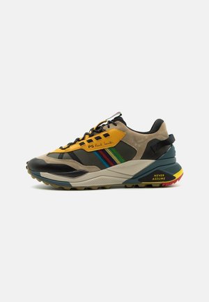 Mehrfarbiger Sneaker mit beigem Wildleder, gelben und schwarzen Details, Regenbogenstreifen an der Seite, klobiger Sohle und dem Schriftzug "Never Assume" an der Ferse.