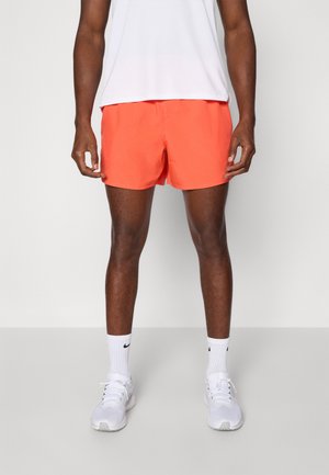 Shorts orange vif, fabriqués en matériau léger. Dotés d'une coupe classique, avec une taille élastique. Portés avec des chaussettes de sport blanches et des chaussures.