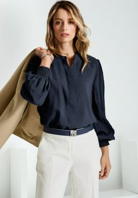 Frau mit blonden Haaren, die eine marineblaue Bluse und weiße Hosen trägt und einen beigen Blazer über die Schulter hält, vor einem schlichten hellen Hintergrund.