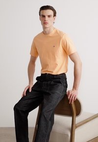 Lys orange bomuld t-shirt med rund hals og lille logo, parret med mørke denimbukser, siddende på en træstol med en blød pude.