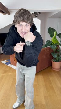 Reloj de pulsera plateado y negro con cara redonda, herrajes metálicos y grandes números. Las características incluyen una correa de goma y sutiles acentos rojos.