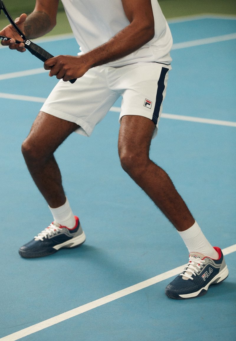 Tenue de tennis pour homme composée d'une chemise blanche et d'un short, avec des accents bleus et rouges. Baskets Fila bleu marine avec des lacets blancs et une semelle en caoutchouc.