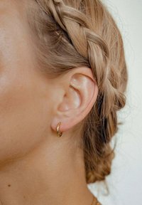 Goldene Creolen, die auf einer Nahaufnahme eines geflochtenen Haarschnitts dargestellt sind; sie haben eine glatte, glänzende Oberfläche und ein kreisförmiges Design.