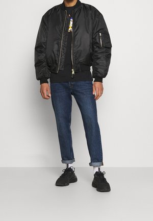 Man met een zwarte bomberjack, een zwarte grafische trui, opgerolde blauwe spijkerbroek, witte sokken en zwarte sneakers, staand tegen een effen achtergrond.