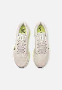 Lys beige treningssko med mesh-overdel, grå snøring, svart Nike-logo og livlig grønn såle. Har en polstret tunge og dempet innersåle.