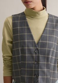 Szara kraciasta kamizelka z dekoltem w serek i trzema guzikami, noszona na jasnooliwkowym golfie. Tkanina o fakturze z kontrastowymi przeszyciami.