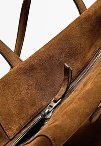 Brown suede handbag with a smooth texture, zip closure, and black leather accents. Interior visible, displaying the zipper pull and lining.