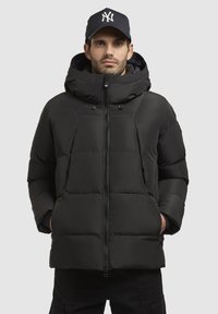 Sort pufferjakke med hætte, med quiltet design, fuld lynlås og snøre detaljer. Båret med en sort kasket.