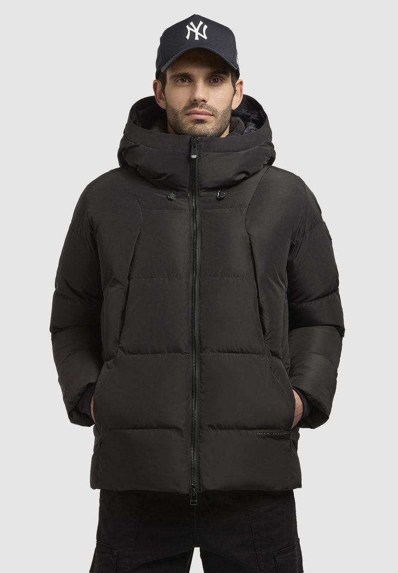 Sort pufferjakke med hætte, med quiltet design, fuld lynlås og snøre detaljer. Båret med en sort kasket.