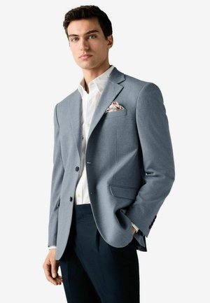 Blazer gris clair avec un revers en pointe, fermeture à un bouton et deux poches avant, porté par-dessus une chemise blanche à boutons et un pantalon sombre.