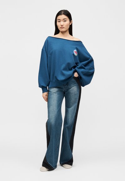 Γυναίκα που φοράει μια φαρδιά μπλε μπλούζα με ακάλυπτους ώμους και φαρδιά τζιν με σκοτεινές πλαϊνές επενδύσεις, στέκεται με το ένα χέρι στην τσέπη.