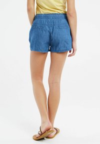 Blå denimshorts med elastisk midja, bakfickor och avslappnad passform, tillsammans med bruna flip-flops.