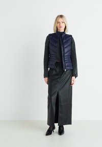 Navy puffer vest with a quilted design, paired with a black knitted sweater and a black leather skirt featuring a front slit. Black shoes.