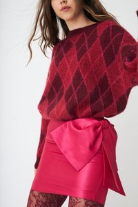 Kobieta ubraną w sweter z czerwono-bordowym wzorem w romby, jasnoróżową spódnicę z dużą kokardą oraz rajstopy w kwiaty, pozująca na białym tle.