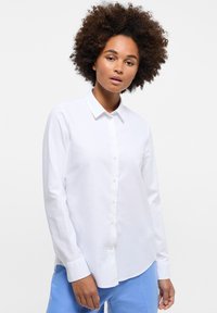 Camicia bianca a maniche lunghe con design a bottoni, caratterizzata da un colletto punta e una consistenza liscia. Indossata con pantaloni azzurri chiari.