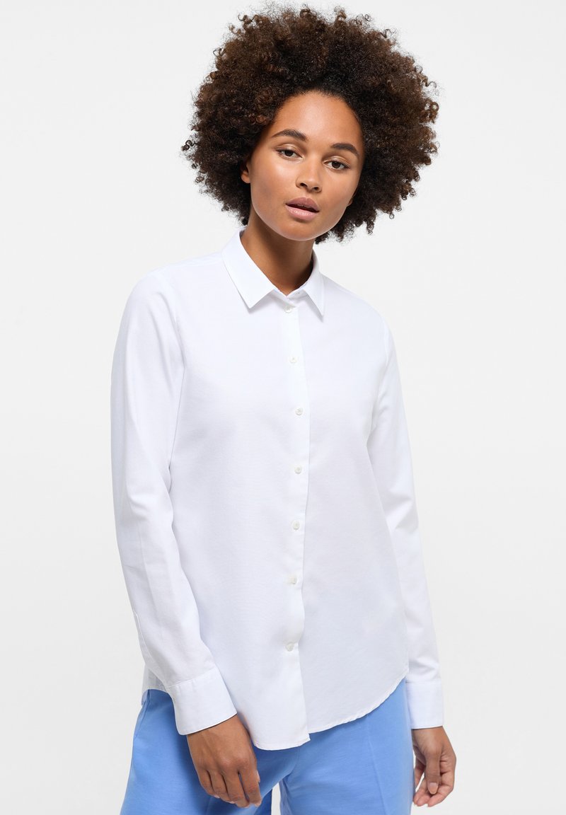 Camicia bianca a maniche lunghe con design a bottoni, caratterizzata da un colletto punta e una consistenza liscia. Indossata con pantaloni azzurri chiari.