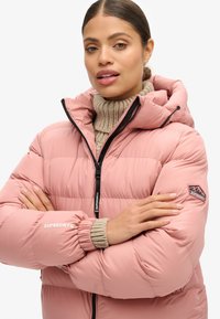Giacca a vento rosa con colletto alto e cappuccio, dotata di una cerniera nera, polsini elastici e un maglione di maglia sotto. Logo Superdry visibile.