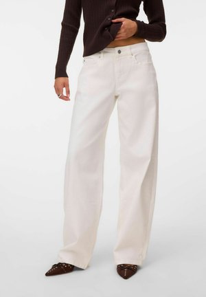 Personne portant un jean blanc taille haute à jambes larges, des chaussures pointues marron, et un pull côtelé marron foncé partiellement relevé pour montrer le ventre.