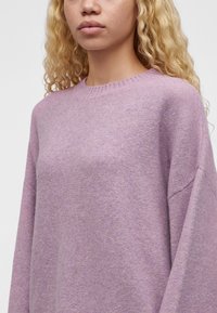 Femme blonde aux cheveux bouclés porte un pull lavande doux et oversize avec un col rond côtelé sur un fond clair uni.