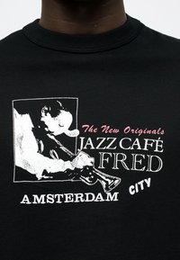Mann spielt Trompete, weißes Grafikdesign mit Text: "The New Originals Jazz Café Fred Amsterdam City" auf schwarzem T-Shirt.