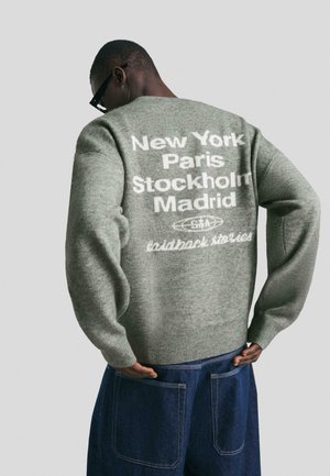 Uomo con un maglione grigio con testo bianco che elenca città e pantaloni blu navy, visto da dietro su uno sfondo semplice.