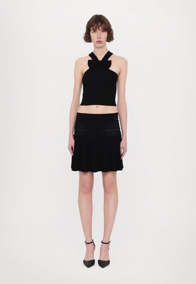 Zwarte gebreide crop top met halternek en strikdetails, gecombineerd met een plissé zwarte rok. Wordt gedragen met pumps met een puntige neus en enkelbandjes.