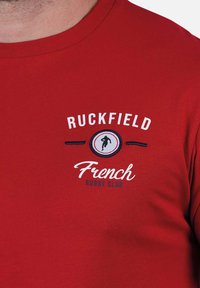 T-shirt en coton rouge portant "Ruckfield French Rugby Club" brodé en bleu et blanc, avec un logo silhouette sportive dans un design circulaire.